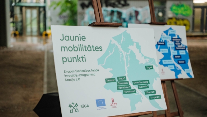 Informatīvs stends uz molberta ar uzrakstu “Jaunie mobilitātes punkti” un Rīgas karti, kurā atzīmētas dzelzceļa staciju apkārtnēs izbūvētās mobilitātes punktu atrašanās vietas.