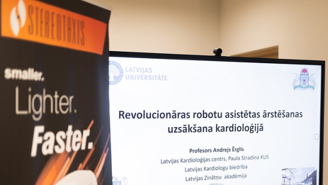 Ekrānā redzama prezentācija “Revolucionāras robotu asistētas ārstēšanas uzsākšana kardioloģijā”; priekšplānā daļēji redzams Stereotaxis stends.