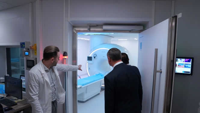 Medicīnas speciālists baltā virsvalkā demonstrē apmeklētājiem magnētiskās rezonanses iekārtu izmeklējumu telpā.
