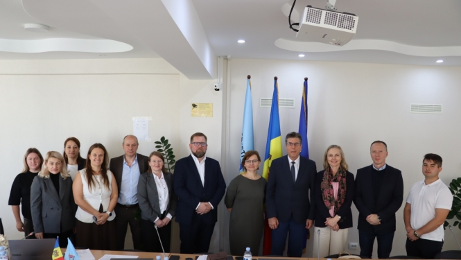 Oficiāls delegācijas kopportrets — 11 cilvēki stāv rindā sanāksmju telpā iepretim valsts karogiem.