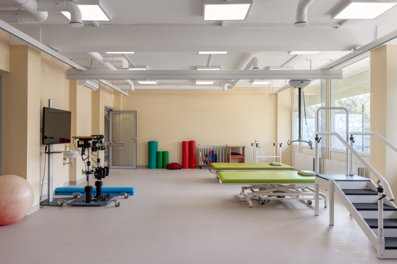 Plaša un gaiša rehabilitācijas telpa ar dažādu fizioterapijas aprīkojumu – vingrošanas bumbu, robotizētu staigāšanas ierīci, paklājiem, krāsainiem putu veltņiem un bumbām pie sienas. Telpā redzamas arī paralēlās stieņi un kāpnes gaitas treniņiem. Lielie logi ielaiž dabisko gaismu, radot patīkamu vidi pacientu rehabilitācijai.
