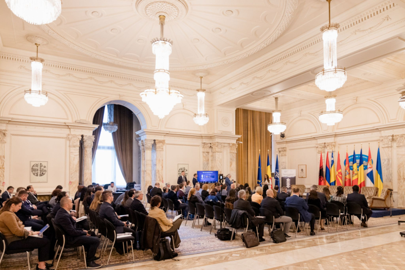 Plaša konferences zāle ar augstiem griestiem un lustru apgaismojumu, kur auditorijai rindās sēž dalībnieki, priekšā skatuve ar prezentāciju ekrānu un vairāku valstu karogiem.