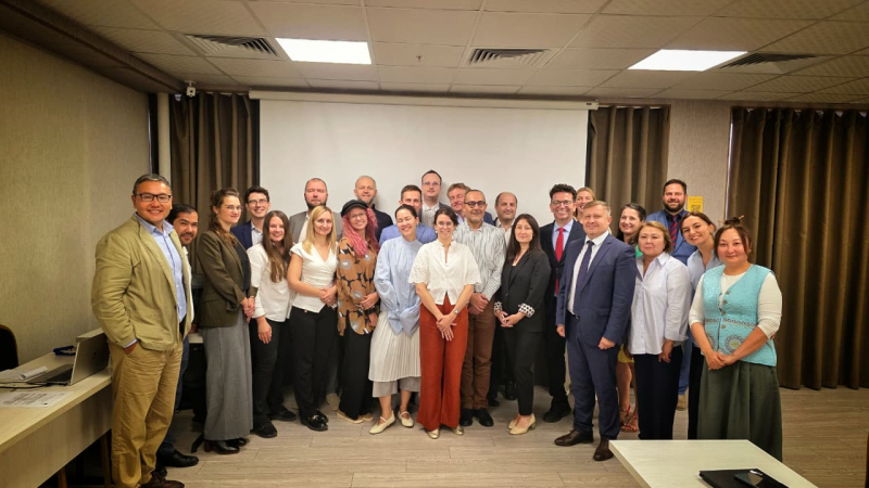 Grupas foto ar apmēram 23 cilvēkiem konferenču telpā, kur redzama balta projekcijas ekrāna siena un brūnas aizkaru dekorācijas fonā. Dalībnieki ir tērpušies biznesa vai ikdienas apģērbā, daļa smaida, citi ar neitrālu izteiksmi. Attēls uzņemts profesionāla pasākuma vai sanāksmes laikā