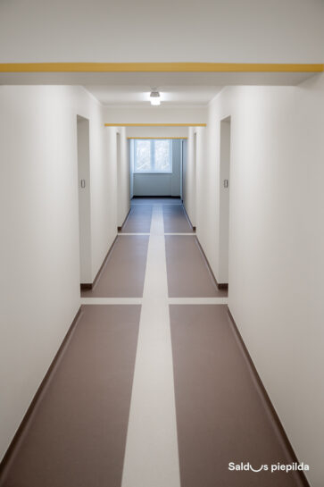 Gaišs, renovēts ēkas koridors ar baltām sienām un brūnu grīdu, kuras vidū ieklāta gaiša, krusta formas līnija; gaiteņa galā redzams logs, durvis izvietotas abās pusēs.
