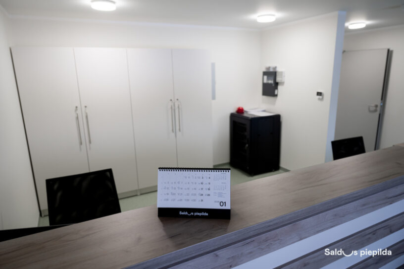 Moderna un gaiša biroja telpa vai pieņemšanas zona ar koka tekstūras leti priekšplānā. Uz letes novietots 2026. gada janvāra galda kalendārs ar uzrakstu "Saldus piepilda". Fonā redzami balti iebūvētie skapji, minimālistisks interjers un melna serveru skapja tipa iekārta pie sienas. Labajā apakšējā stūrī ir logotips "Saldus piepilda".