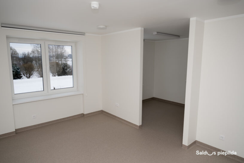 Gaiša, tukša dzīvojamā telpa ar pelēku grīdu un baltām sienām; kreisajā pusē liels logs ar skatu uz sniegotu ainavu, telpa sadalīta divās zonās ar starpsienu.