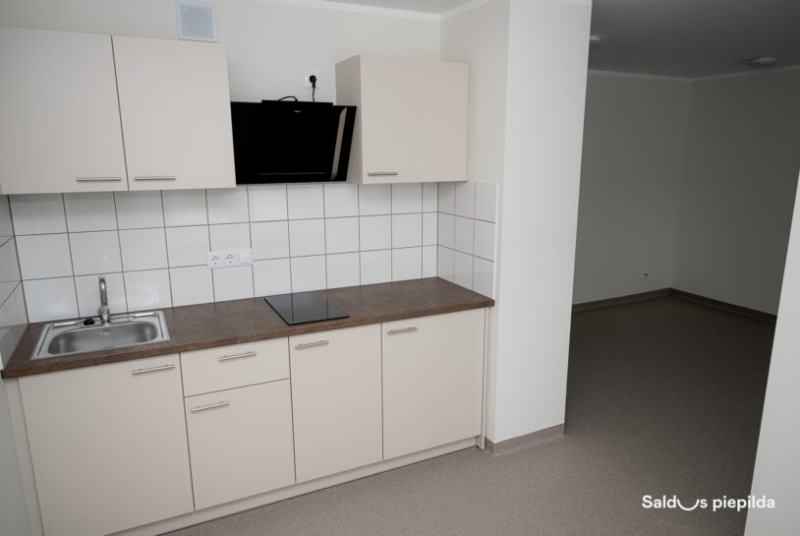 Neliela, gaiša virtuves zona ar gaišiem skapīšiem, izlietni, elektrisko plīti un nosūcēju; sienas apdarē balti flīžu paneļi, blakus atrodas tukša dzīvojamā telpa ar pelēku grīdu.