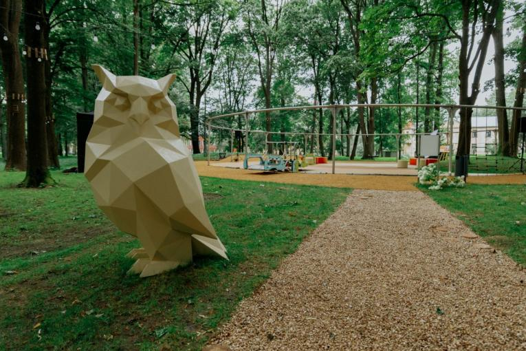 Parks ar lielu, ģeometrisku pūces skulptūru kreisajā pusē, veidotu no gaiša materiāla ar šķautņainām formām. Fonā redzama rotaļu zona ar šūpolēm un kāpšanas konstrukcijām, apkārt aug gari koki, un priekšplānā ved grants celiņš uz rotaļu laukumu.