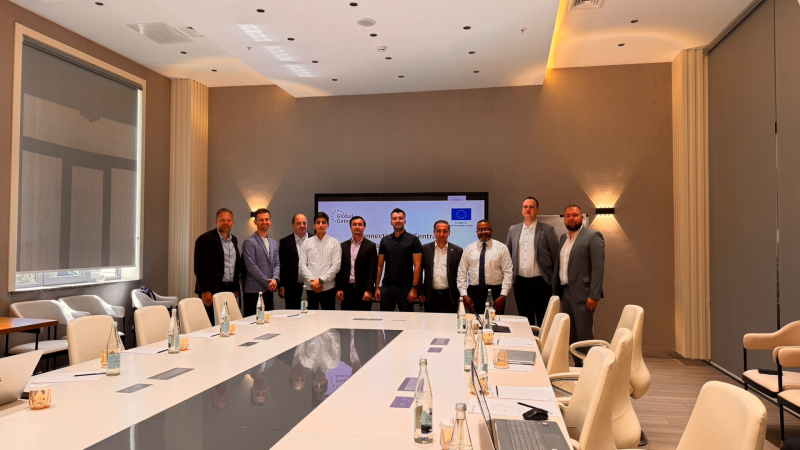 Desmit cilvēku grupa konferenču telpā stāv aiz garas galda, uz kura redzamas ūdens pudeles, dokumenti un klēpjdatori. Fonā liels ekrāns ar “Global Gateway” un Eiropas Savienības logotipiem, kā arī uzrakstu “Connecting Central Asia”. Telpa ar modernu interjeru – bēšas sienas un griestu apgaismojums