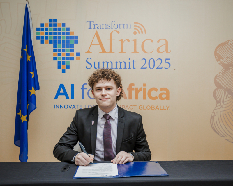 Persona sēž pie galda un paraksta dokumentu, fonā redzams plakāts ar uzrakstu “Transform Africa Summit 2025” un “AI for Africa – Innovate Locally, Impact Globally”, kā arī Eiropas Savienības karogs kreisajā pusē.