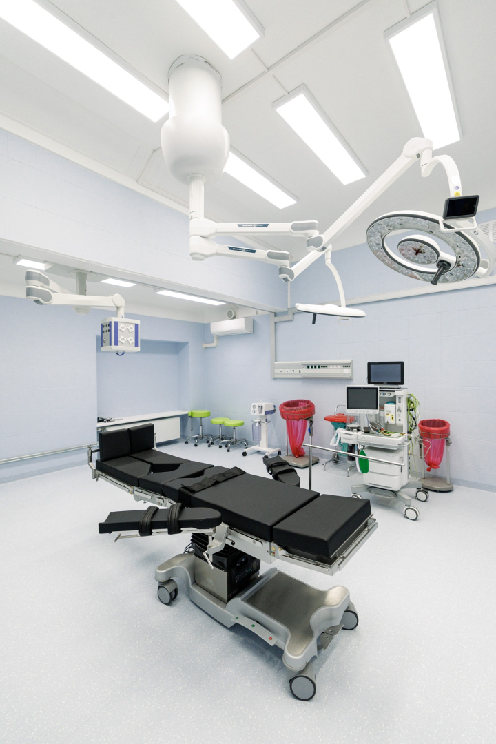 Mūsdienīga operāciju zāle ar regulējamu ķirurģisko galdu centrā, ko ieskauj medicīniskā aparatūra un monitori. Griestos redzamas ķirurģiskās lampas un iekārtu turētāji. Telpas sienas gaiši zilas, grīda balta un tīra; fonā redzami sarkani atkritumu konteineri un zaļi krēsli.