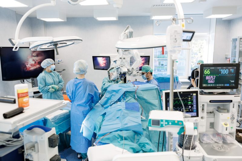 Mūsdienīga operāciju zāle ar ķirurgu komandu, kas veic operāciju. Telpā redzama augstas klases medicīniskā aparatūra – monitori ar ķirurģiskiem attēliem, anestēzijas iekārta un citi medicīnas instrumenti. Ķirurgu komanda tērpta sterilās halātēs, maskās un cimdiem; pacients atrodas uz operāciju galda, pārklāts ar zilām steriliem pārklājiem.