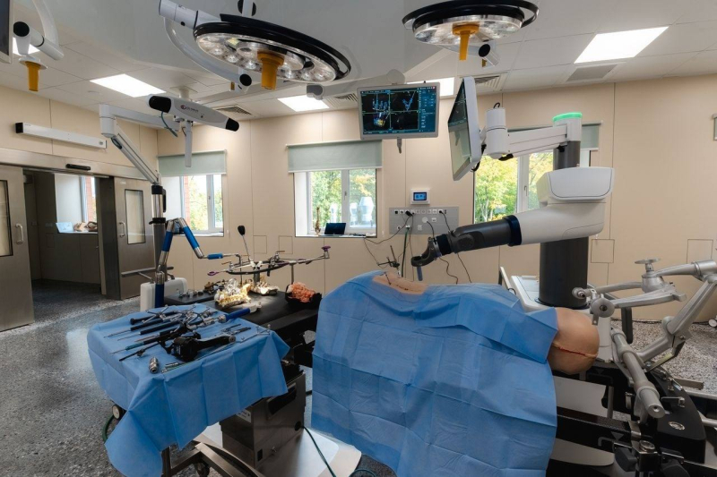 Moderna operāciju zāle ar augstas precizitātes medicīnisko aprīkojumu – virs operāciju galda ar zilām steriliem pārklājiem novietotas robotizētas ķirurģiskās rokas, blakus galds ar ķirurģiskajiem instrumentiem, griestos – apgaismojums un monitori ar medicīniskiem attēliem. Telpā ir lieli logi, kas ielaiž dienasgaismu, un kopējā vide ir tīra un sakārtota, gatava operācijai