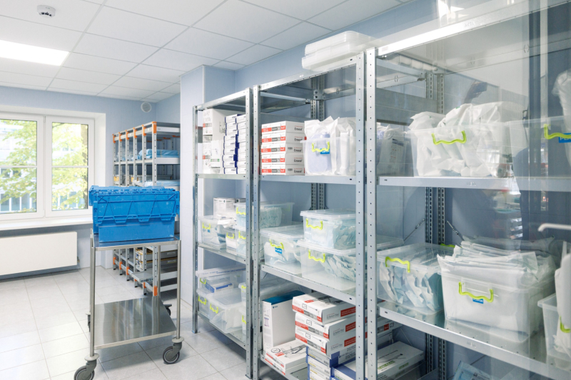 Sakārtota noliktavas telpa medicīnas iestādē ar metāla plauktiem, kuros glabājas medicīniskie piederumi kastēs un caurspīdīgos konteineros ar dzeltenām etiķetēm; priekšplānā redzams zils plastmasas konteiners uz ratiņiem. Telpa ir gaiša un tīra, ar lieliem logiem, kas ielaiž dienasgaismu.