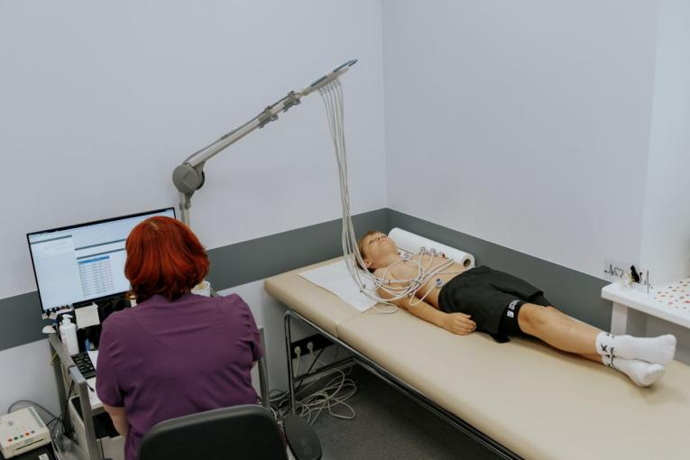 Medicīnas darbiniece ar sarkanīgiem matiem sēž pie datora galda un novēro pacientu, kurš guļ uz izmeklējumu galda. Pacientam, jaunam cilvēkam, pie krūtīm un ekstremitātēm ir pievienoti vairāki elektrodi, kas ar vadiem savienoti ar medicīnisko aparatūru. Telpa izskatās pēc klīniskās vides ar redzamu medicīnisko aprīkojumu.