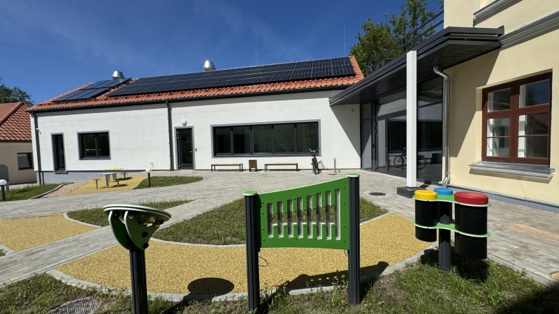 Mūsdienīga ēka ar saules paneļiem uz sarkaniem dakstiņu jumta, baltu fasādi un lieliem logiem. Priekšplānā redzama bērnu rotaļu zona ar dzeltenu gumijotu segumu, interaktīviem rotaļu elementiem, tostarp krāsainiem bungām un zaļu paneli ar vertikālām spraugām. Apkārt ir bruģēti celiņi, un viss ir izgaismots saulainā dienā zem skaidrām zilām debesīm.