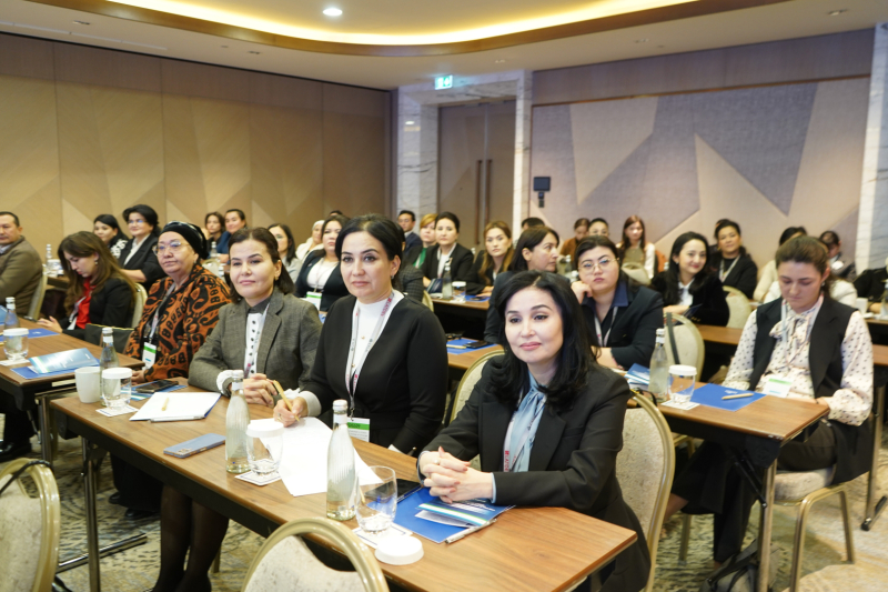 Konferences dalībnieki sēž zālē pie galdiem, klausoties prezentāciju; uz galdiem redzami bloki, pildspalvas, ūdens pudeles un kafijas tases.