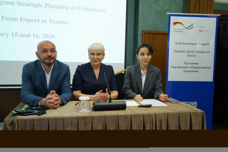 Trīs cilvēki sēž pie galda preses vai semināra situācijā, priekšā mikrofonam un dokumentiem. Aiz viņiem redzams prezentācijas ekrāns ar informāciju par stratēģiskās plānošanas programmu Uzbekistānā, bet blakus – stends ar Good Governance Program nosaukumu vairākās valodās.