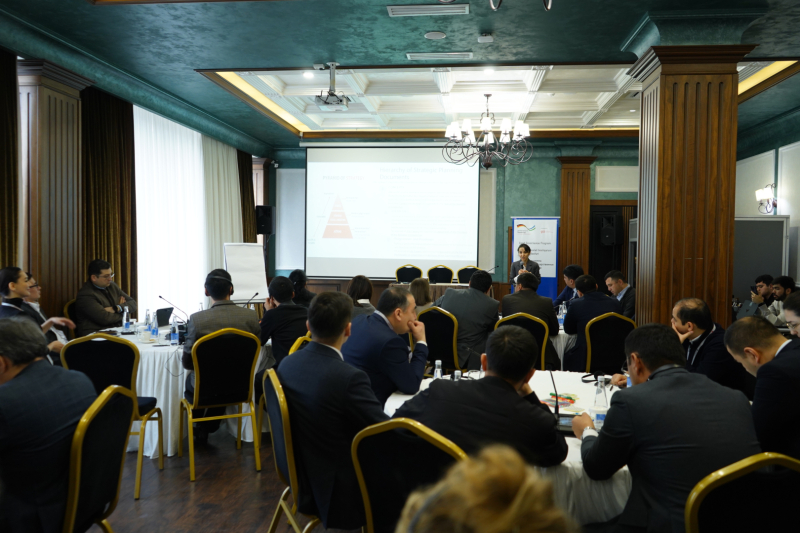 Konferenču zāle ar apaļiem galdiem, pie kuriem sēž dalībnieki formālā apģērbā; priekšā redzama prezentācija uz ekrāna un lektore pie tribīnes, fonā – pasākuma baneris un lustra.