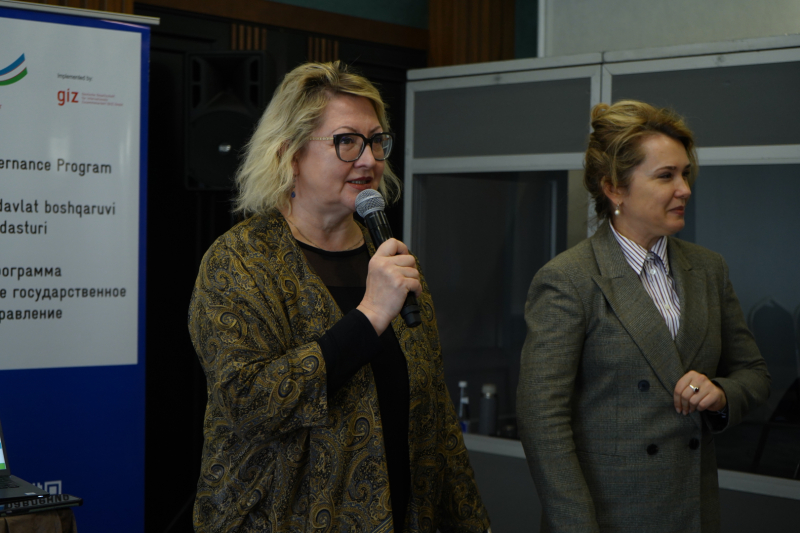 Divas sievietes stāv pasākuma telpā; viena runā mikrofonā, otra stāv blakus un klausās. Fonā redzams informatīvs baneris un konferenču aprīkojums.