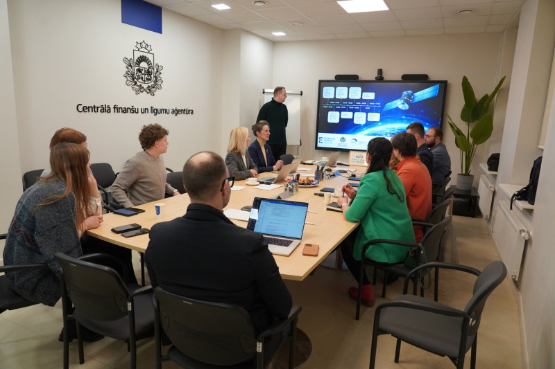CFLA telpās notiek darba sanāksme – pie galda sēž projekta partneri, priekšā redzams ekrāns ar prezentāciju par satelītu risinājumiem un digitālo savienojamību