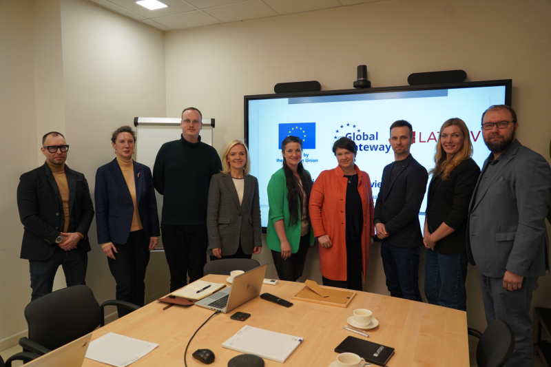 Latvijas partneru projekta atklāšanas pasākuma dalībnieki stāv kopbildē CFLA telpās, fonā redzams ES “Global Gateway” iniciatīvas vizuālais noformējums.