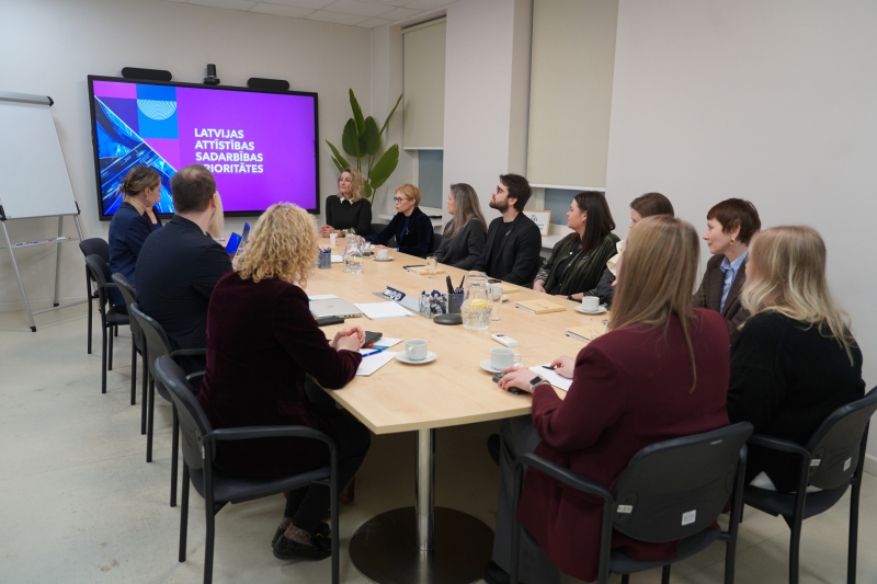 Liela darba sanāksme ap konferenču galdu, kur dalībnieki klausās prezentāciju par Latvijas attīstības sadarbības prioritātēm. Fonā redzams liels ekrāns ar prezentācijas slaidu, uz galda – dokumenti, klades, kafijas tasītes un rakstāmpiederumi.