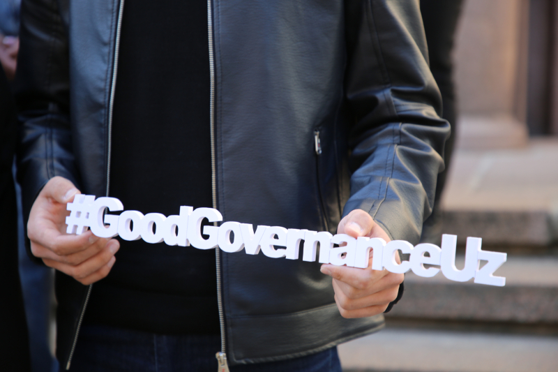 Tuvplāna foto ar cilvēku, kas stāv uz āra kāpnēm un rokās tur baltu uzrakstu “#GoodGovernanceUz”.