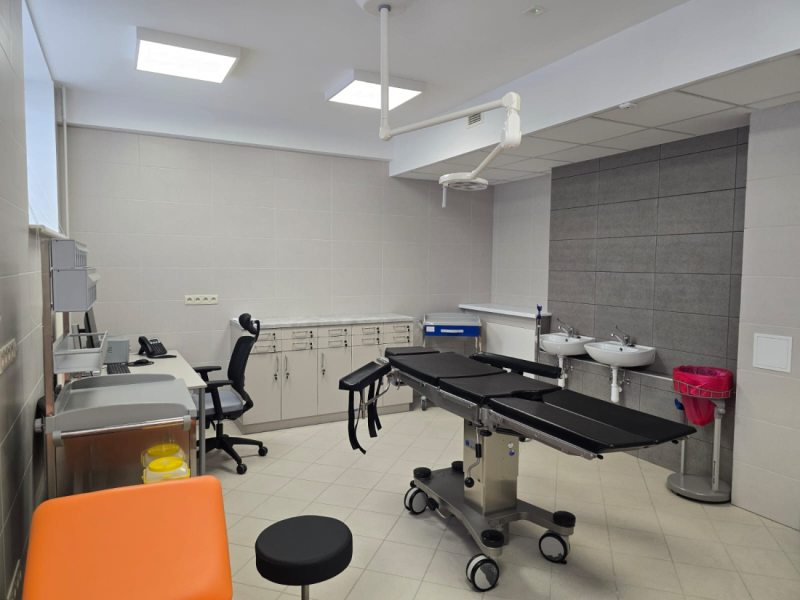 Moderni aprīkota ārstniecības telpa ar regulējamu operāciju galdu telpas centrā, griestos stiprinātu ķirurģisko apgaismojumu, izlietnēm, skapīšiem instrumentiem un darba vietu ar datoru; telpa ir gaiša, sterila un funkcionāla