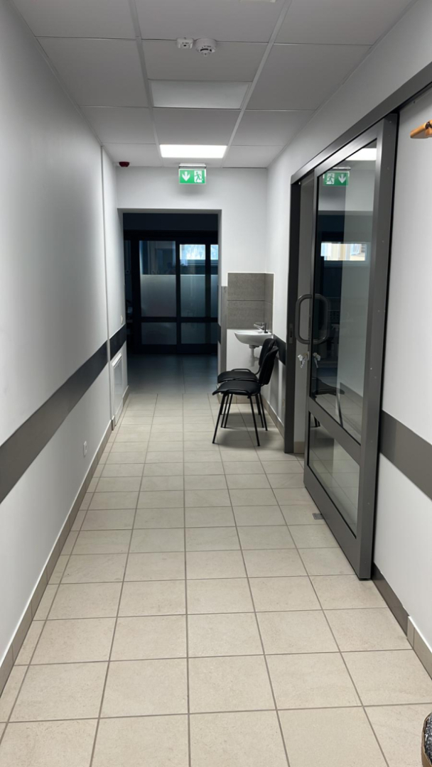 Gaišs medicīnas iestādes gaitenis ar flīzētu grīdu, stiklotām durvīm labajā pusē, izlietni un saliktiem krēsliem gaidīšanai; griestos redzama avārijas izejas norāde, telpa ir tīra un funkcionāla.