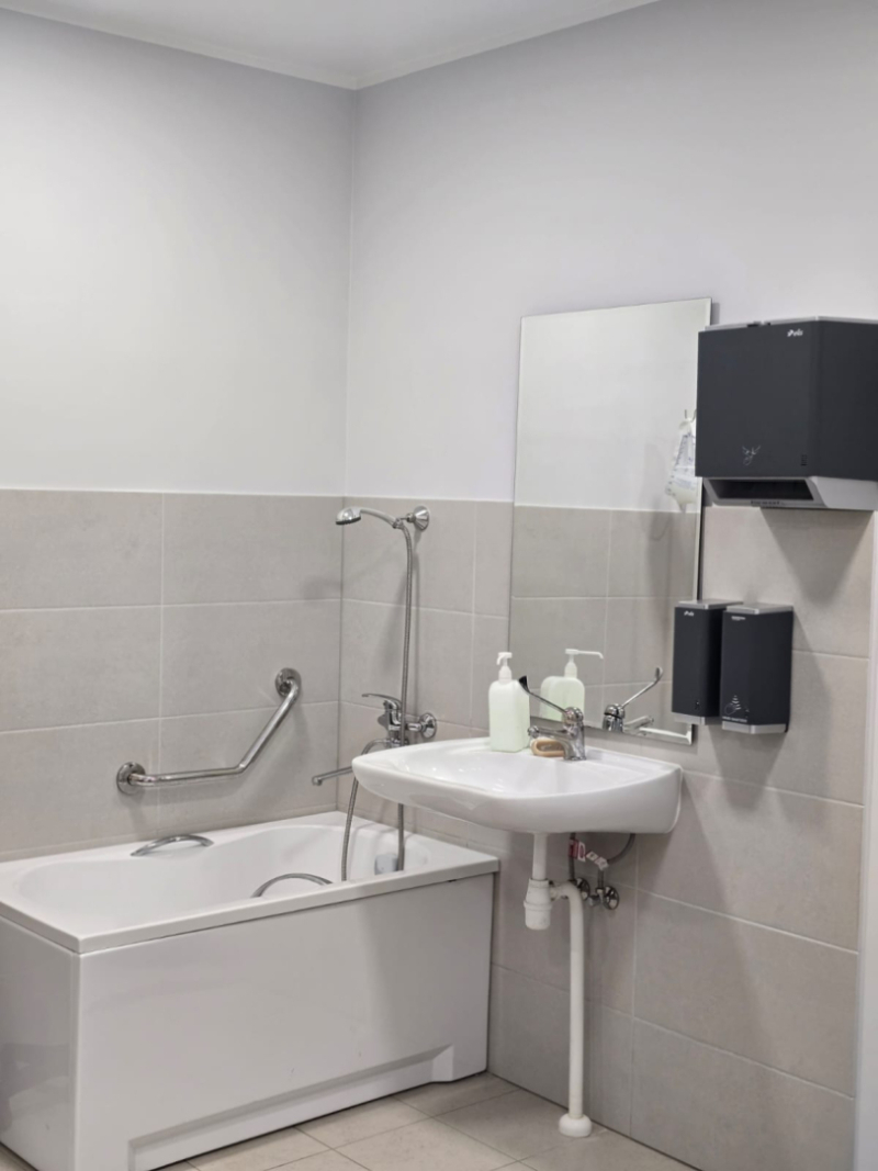 Vannasistaba medicīnas iestādē ar pielāgotu vannu un drošības rokturi, izlietni ar spoguli, ziepju un papīra roku dvieļu dozatoriem; telpa ir gaiša, tīra un piemērota pacientu higiēnas vajadzībām.