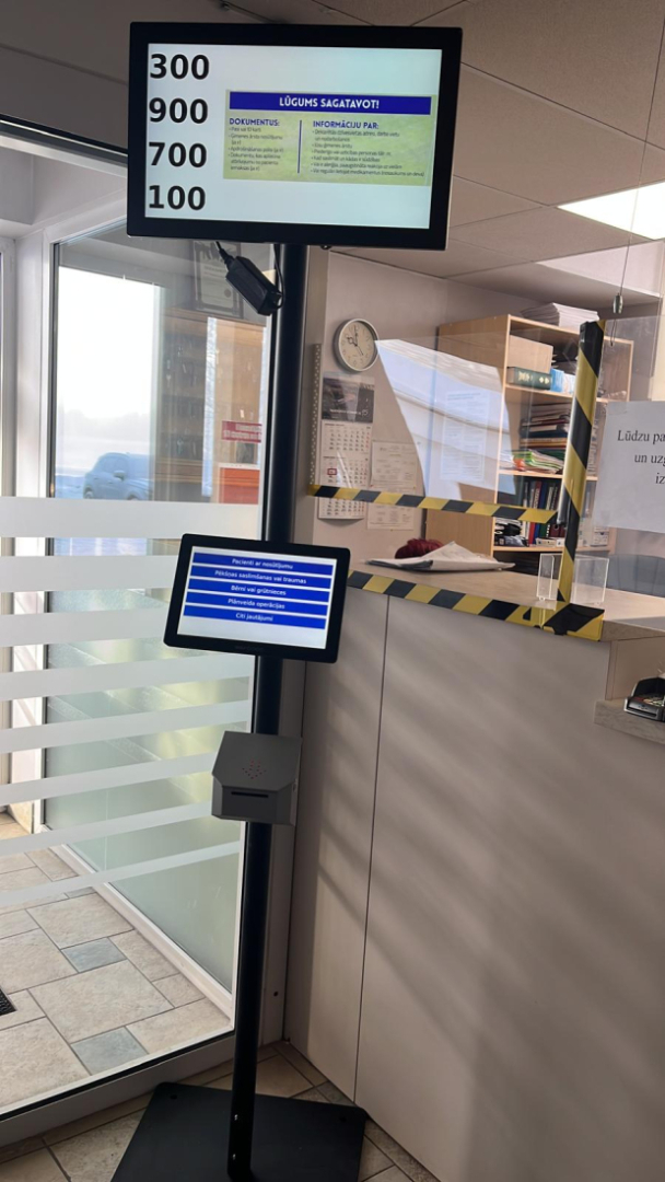 Pacientu apkalpošanas zona ar elektronisko rindas vadības stendu: uz statīva izvietoti divi ekrāni, augšējā redzami apkalpošanas numuri, apakšējā – izvēlnes pakalpojumu veidiem; fonā stiklotas durvis un reģistratūras lete ar aizsargstiklu.
