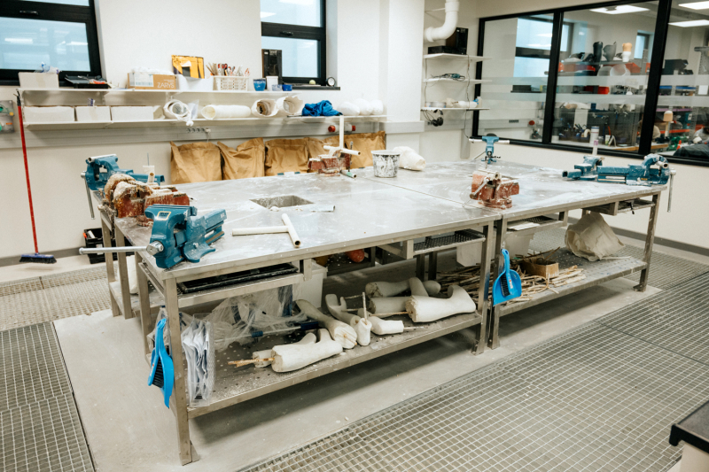 Darbnīcas telpa ar lielu darba galdu, skavām, instrumentiem, materiāliem un plauktiem pie sienas.