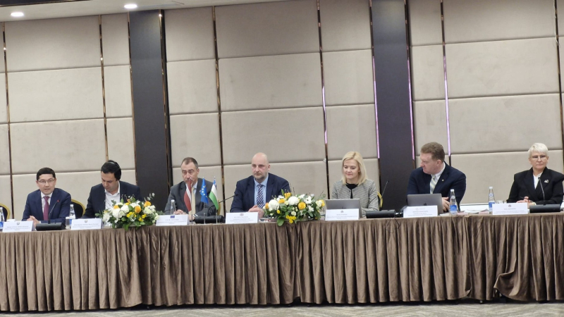 Konferenču zālē pie gara galda sēž seši amatpersonas – četri vīrieši un divas sievietes – oficiāla projekta atklāšanas pasākumā. Uz galda novietotas ziedu kompozīcijas, ūdens pudeles, mikrofoni un vārda kartītes, kā arī redzami Eiropas Savienības un Uzbekistānas karodziņi. Dalībnieki ir tērpti lietišķā apģērbā un piedalās paneļdiskusijā vai preses konferencē.