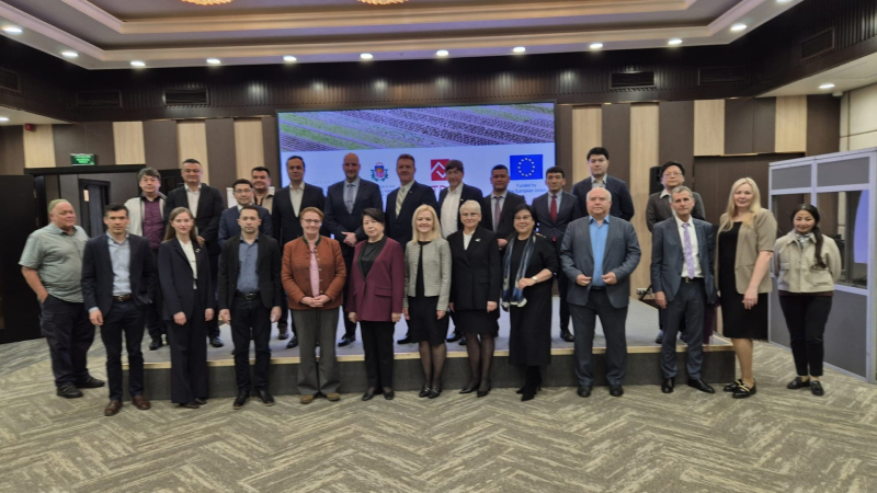 Konferenču zālē uz skatuves divās rindās pozē apmēram divdesmit pieci pasākuma dalībnieki – vīrieši un sievietes lietišķā apģērbā. Aiz viņiem redzams ekrāns ar projekta vizuālo materiālu un Eiropas Savienības karogu. Fotogrāfija uzņemta pēc oficiāla pasākuma vai projekta atklāšanas.