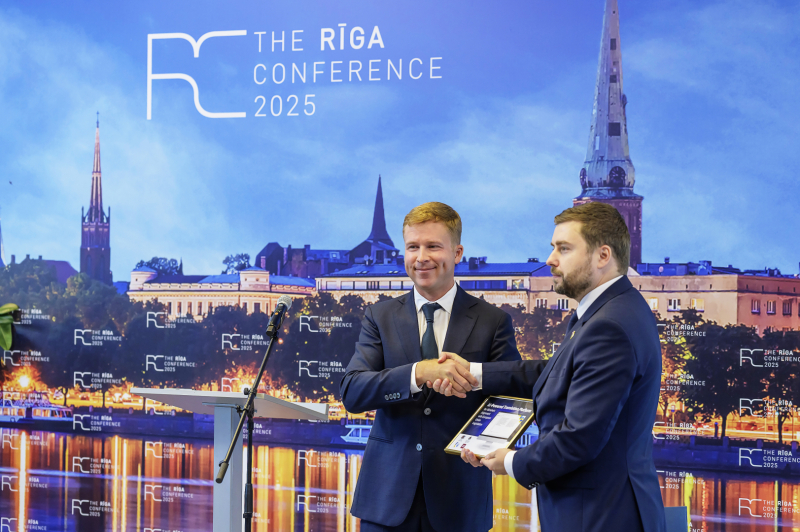 Divi cilvēki formālā apģērbā paspiež roku uz skatuves ar Rīgas panorāmu fonā, notiek apbalvošanas ceremonija 'The Rīga Conference 2025' ietvaros.
