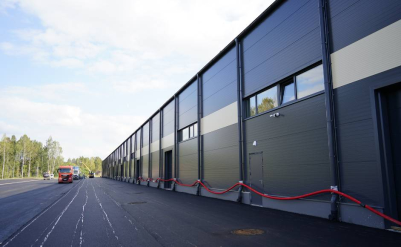 Liela industriāla ēka ar garu, tumši pelēku fasādi, kurā izvietoti logi un durvis. Gar sienas apakšdaļu horizontāli novietotas sarkanas šļūtenes. Fonā redzams sarkans kravas auto uz asfalta ceļa, kā arī koki un citas automašīnas. Debesis daļēji mākoņainas.