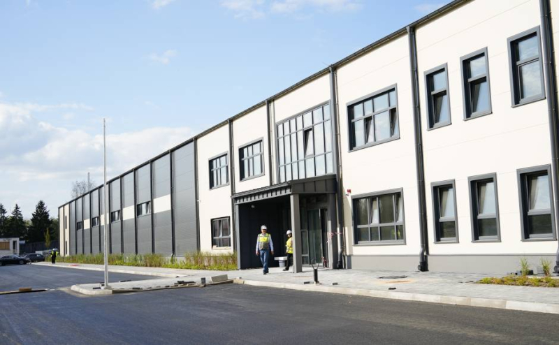 Mūsdienīga divstāvu industriāla vai komerciāla ēka ar baltu un tumši pelēku fasādi, lieliem logiem un galveno ieeju ar nelielu nojumi. Priekšpusē redzami divi cilvēki drošības vestēs un ķiverēs, kas dodas uz ēku. Apkārtne ir labiekārtota ar jauniem ceļiem, ietvēm un stādījumiem. Debesis ir skaidras ar dažiem mākoņiem.