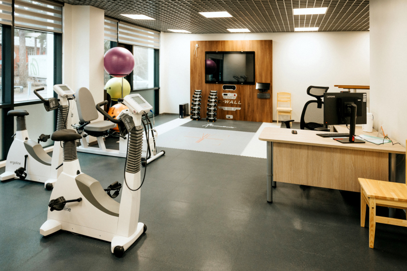 Fizioterapijas telpa ar trenažieriem, velotrenažieriem, vingrošanas bumbām, darba galdu un sienas ekrānu.