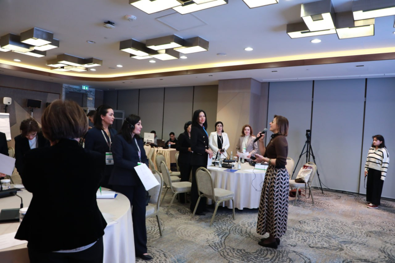 Diskusijas brīdis seminārā konferenču zālē; dalībnieki stāv pie galdiem, viena persona runā mikrofonā, fonā redzama prezentācijas tehnika un kameras.