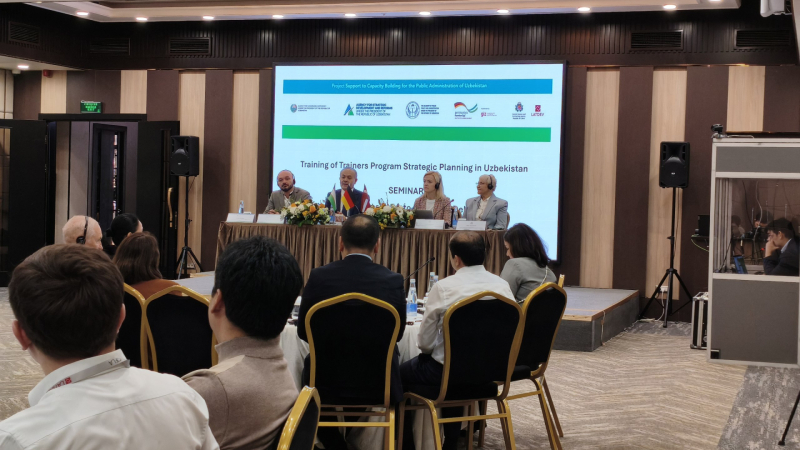 Četri eksperti, tostarp no Latvijas, sēž pie diskusiju galda, kas rotāts ar ziediem un galda karodziņiem. Uz lielā ekrāna aiz viņiem redzams uzraksts "Training of Trainers Program Strategic Planning in Uzbekistan". Priekšplānā redzami semināra dalībnieki no mugurpuses, klausoties prezentāciju.