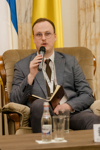 Persona sēž krēslā un runā mikrofonā, rokās turot piezīmju bloku; fonā redzami vertikāli izvietoti karogi.