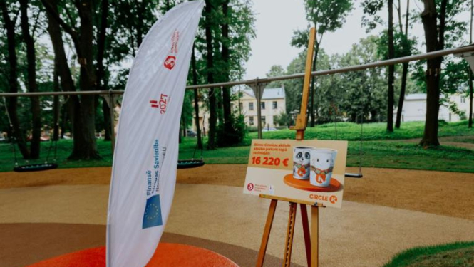 Āra teritorija ar rotaļu laukumu fonā. Priekšplānā redzama balta karogs ar tekstu un logotipiem, kā arī molberts ar plakātu, uz kura attēloti divi bundžu attēli un teksts: "Bērnu pilsētas aktivitāšu centra izveidei piešķirts līdzfinansējums 16 220 €".
