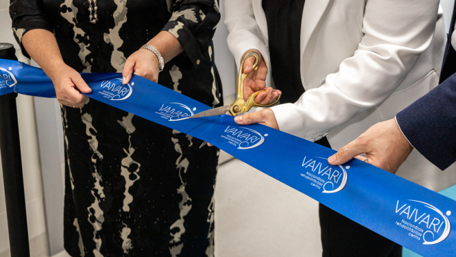 Trīs cilvēki piedalās lentes griešanas ceremonijā pie VAIVARI Nacionālā rehabilitācijas centra. Viens no viņiem ar lielām zelta šķērēm pārgriež zilu lenti ar centra nosaukumu, atzīmējot jaunas ēkas vai pakalpojuma atklāšanu.