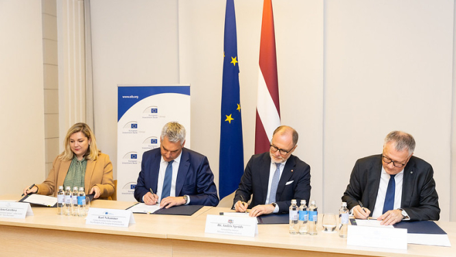 4 pārstāvji paraksta dokumentus pie galda, fonā redzamas Eiropas Savienības un Latvijas karogi, kā arī Eiropas Investīciju bankas (EIB) baneris ar vietni www.eib.org. Attēls no oficiālas parakstīšanas ceremonijas.