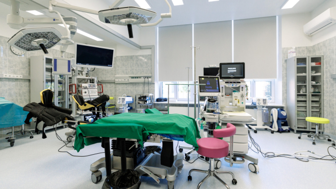 Mūsdienīga operāciju zāle ar centrāli novietotu operāciju galdu, kas pārklāts ar zaļu pārklāju. Telpā redzama medicīniskā aparatūra – ķirurģiskās lampas, monitori, anestēzijas iekārta un citi instrumenti. Fonā redzami plaukti un skapji ar medicīnas piederumiem, kā arī logi ar daļēji aizvilktām žalūzijām. Priekšplānā – rozā krēsls.