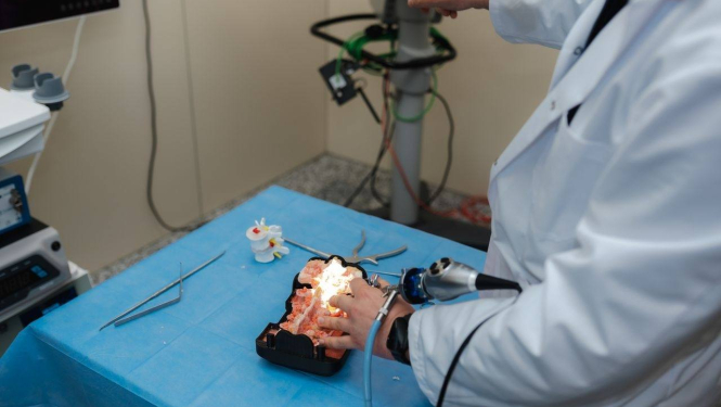 Persona baltā halātā veic manipulācijas ar medicīnisku vai anatomisku galvas/galvas smadzeņu modeli, kas novietots uz zilas ķirurģiskās drānas. Tiek izmantoti medicīnas instrumenti, tostarp endoskops vai tam līdzīga ierīce. Apkārt redzami dažādi rīki un aprīkojums, norādot uz medicīnas laboratoriju vai apmācību telpu, kur notiek praktiska procedūra vai mācību sesija.