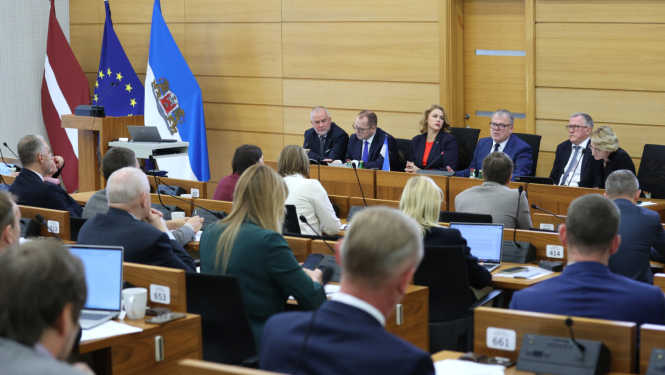 Attēlā redzama sanāksme, kurā piedalās vairāki cilvēki, sēžot pie galda un klausoties prezentāciju vai diskusiju. Priekšplānā ir vairāki cilvēki ar klēpjdatoriem un dokumentiem. Fonā ir Latvijas karogs, Eiropas Savienības karogs un vēl viens karogs ar ģerboni.