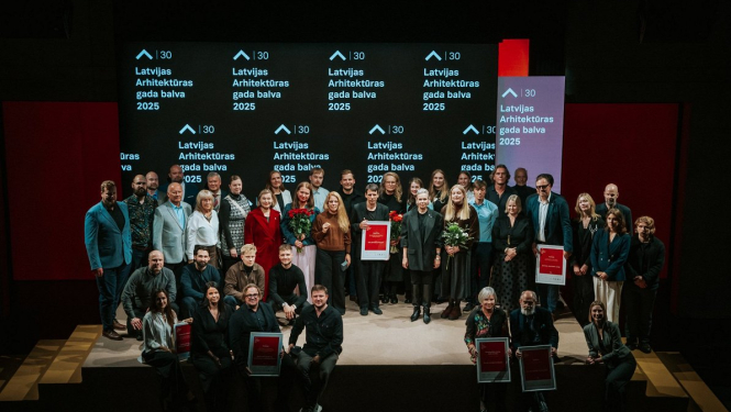 Grupas fotogrāfija ar cilvēkiem, kas stāv uz skatuves un tur rokās balvas. Fonā redzams ekrāns ar uzrakstu "Latvijas Arhitektūras gada balva 2025"