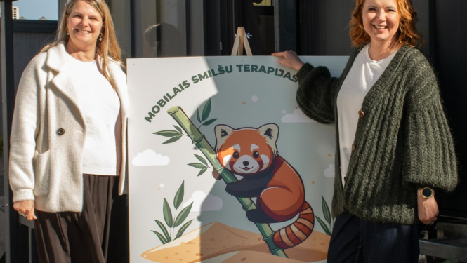 Divi cilvēki stāv pie plakāta ar ilustrāciju, kurā redzama sarkana panda ar bambusu. Uz plakāta rakstīts 'Mobilais smilšu terapijas'. Fonā redzami augi un smilšu kāpas, radot dabas un terapijas noskaņu